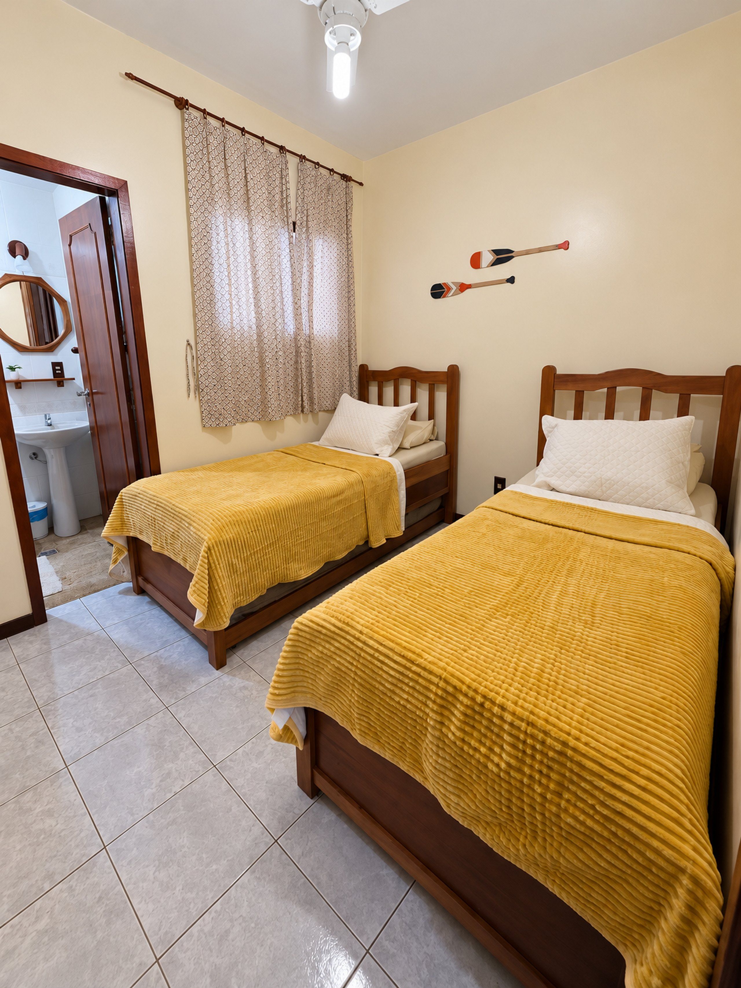 A room with two wooden beds, a bathroom with a mirror, and a ceiling fan.