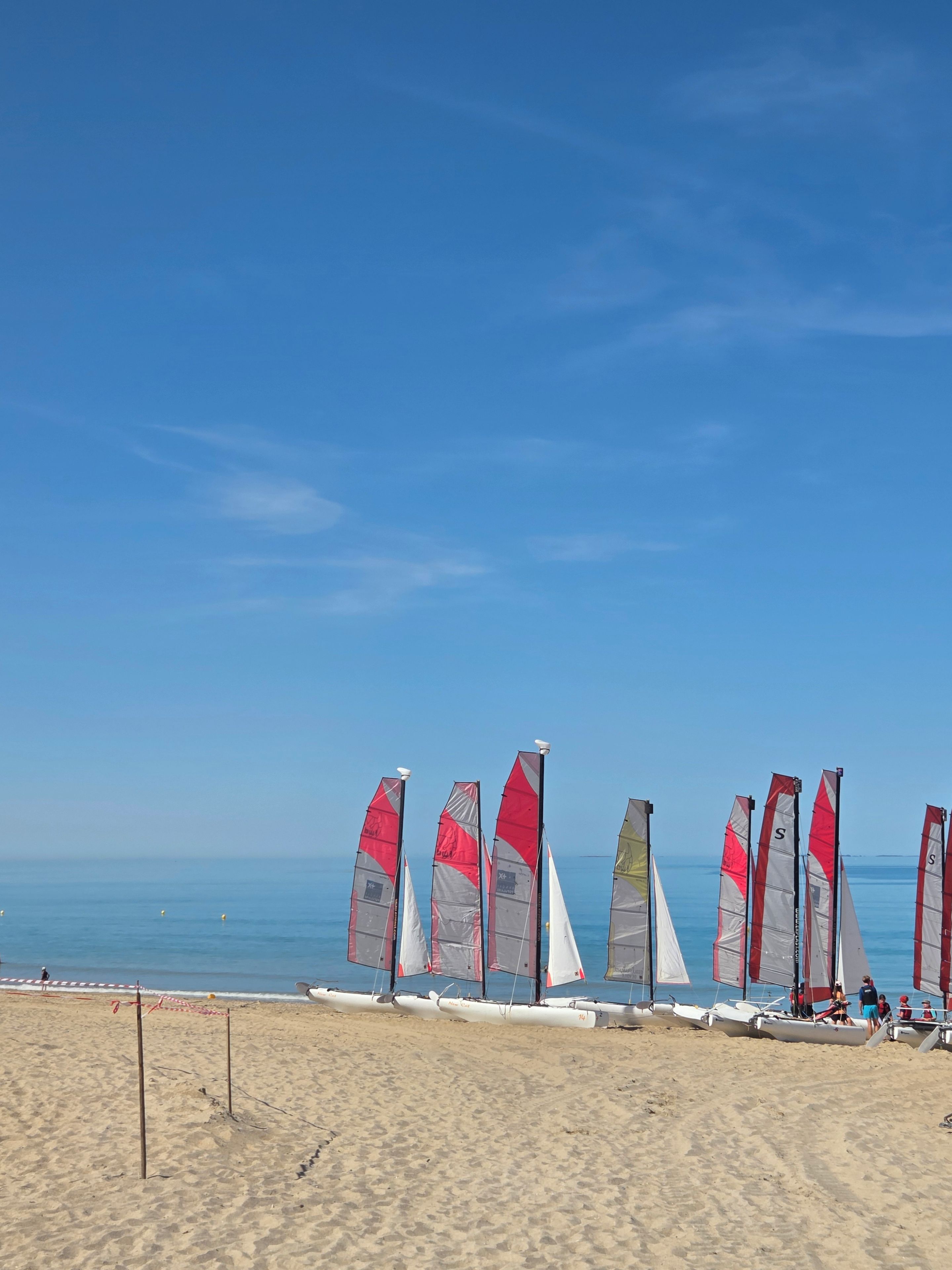 Uma praia com vários veleiros enfileirados na areia, céu claro e oceano ao fundo.