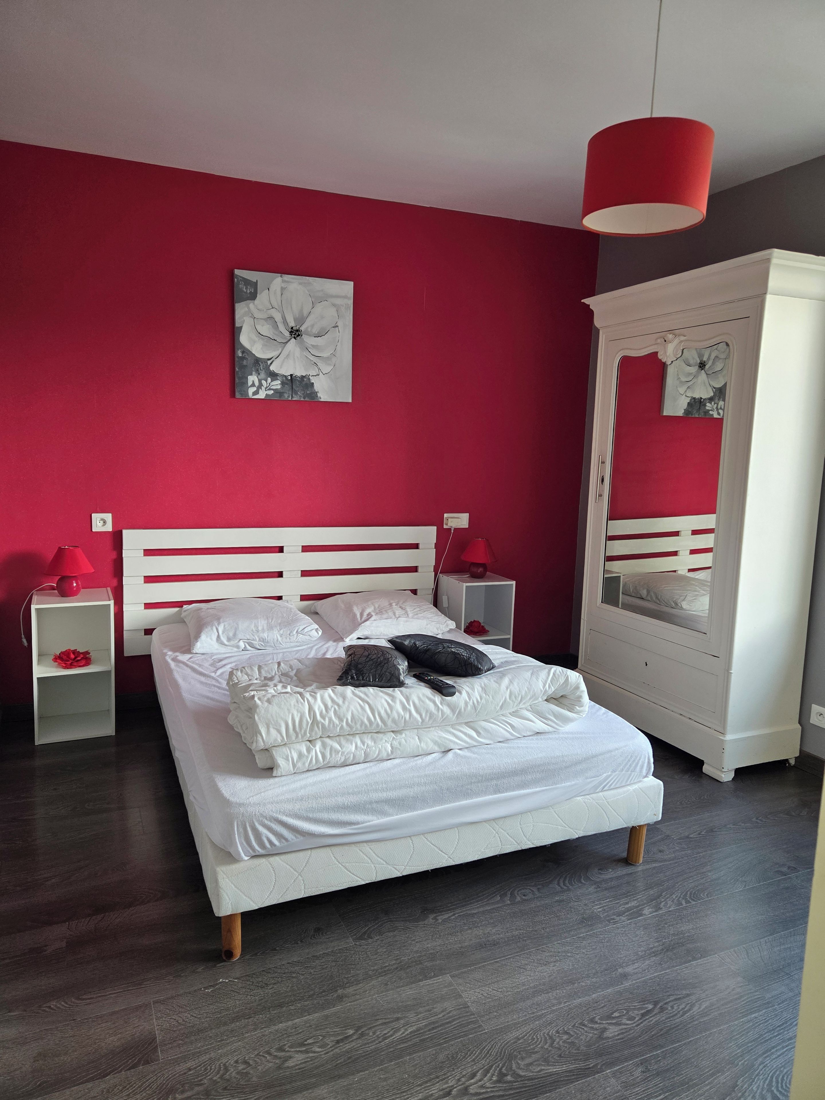 Une chambre avec un lit blanc, des murs rouges, un luminaire suspendu rouge, une table de chevet blanche et une armoire blanche avec un miroir.