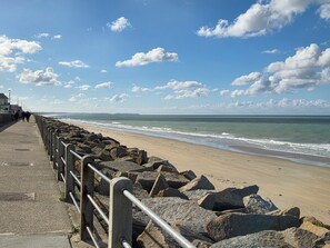 Une promenade en bord de mer, dotée d’une rambarde métallique, qui mène à une plage de sable.