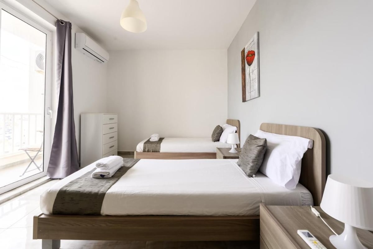 Et hotelværelse med to senge, et natbord med lampe, et garderobeskab og et vindue med gardiner.