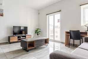 A modern living room with a flat-screen TV, a wooden cabinet, a grey sofa, a desk with a chair, and a large window with a view of buildings. - ATLANTIC COURT F10 (Msida)
