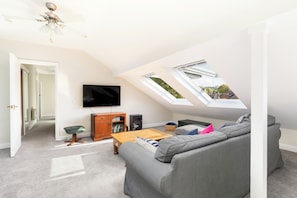 A spacious loft bedroom with a large window, a ceiling fan, a flat-screen TV, a wooden cabinet, and a grey sofa. - Charming Historic 2 Bed & Parking|Stroud Hill View - Pass the Keys (Stroud)