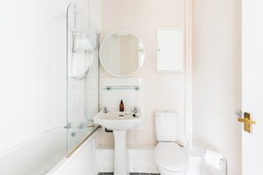 Et badeværelse med glasbrusekabine, hvid vask, toilet og spejl.