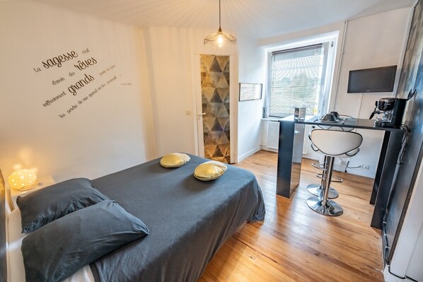 Quarto com cama, travesseiros, abajur, parede com frase inspiradora, porta com padrão geométrico e cozinha compacta com pia e televisão.