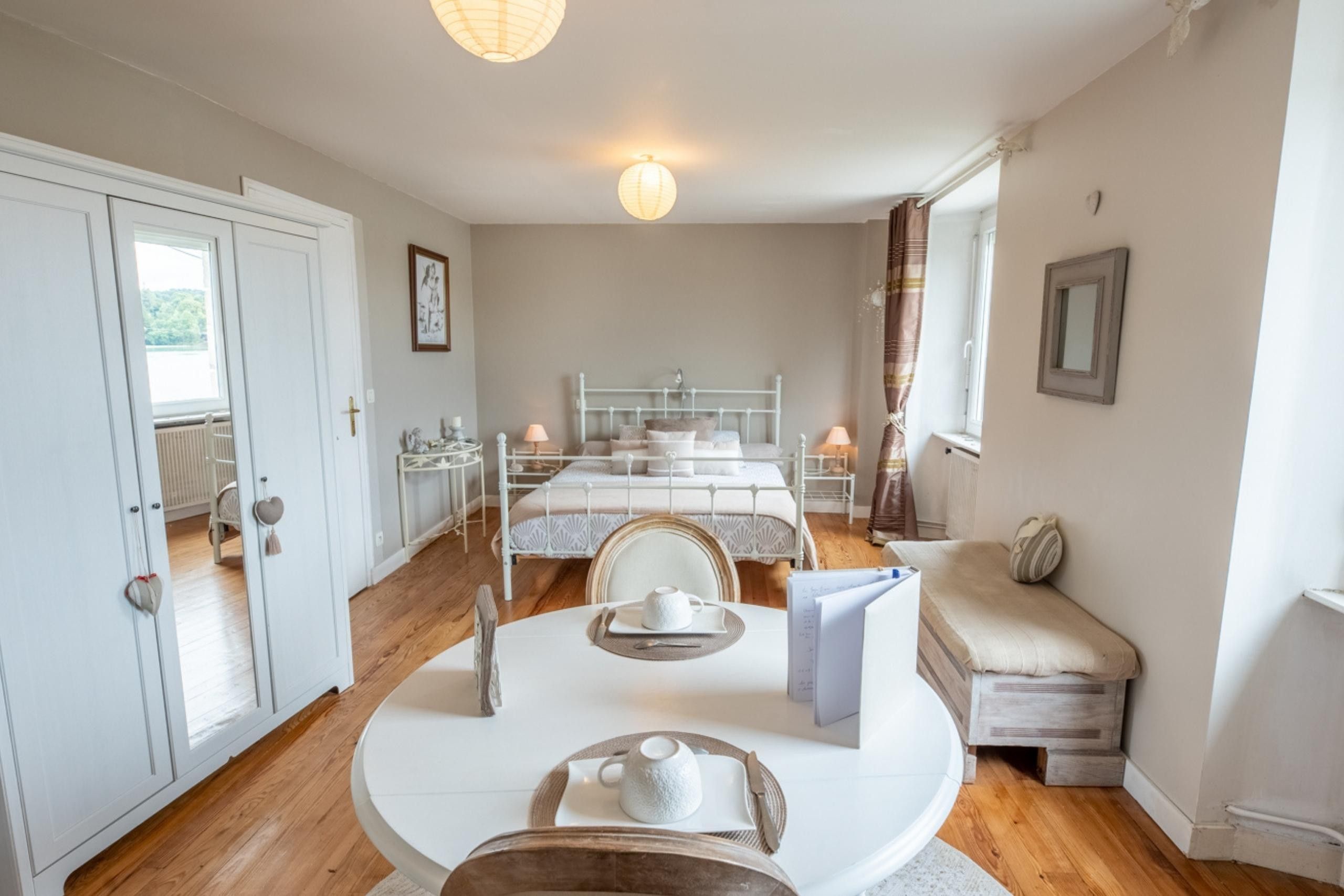A bedroom with a double bed, bedside tables, a chair, a table with a teapot, and a window with curtains.