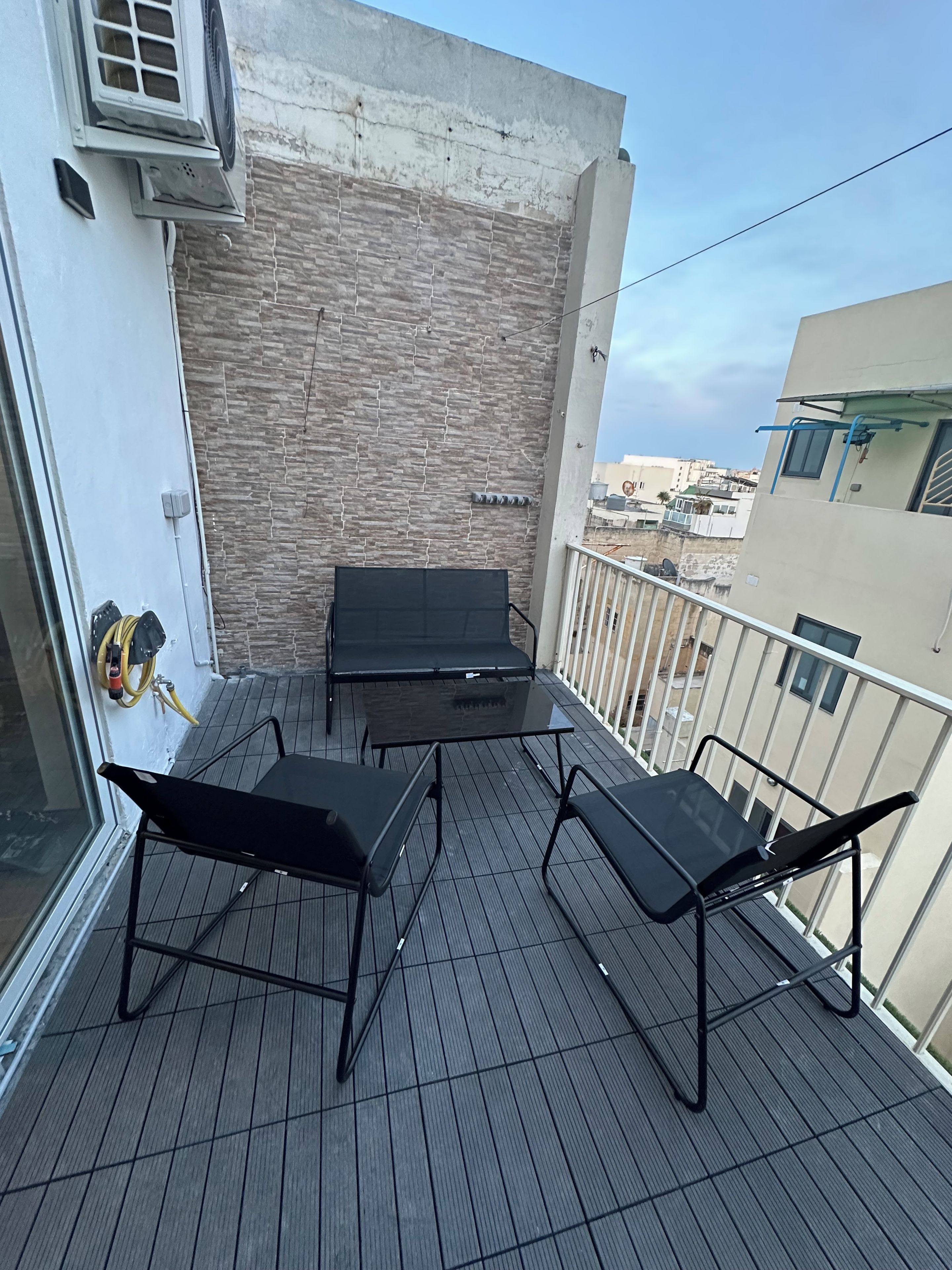 A balcony with a black metal table and chairs, a stone wall, and a view of the cityscape.