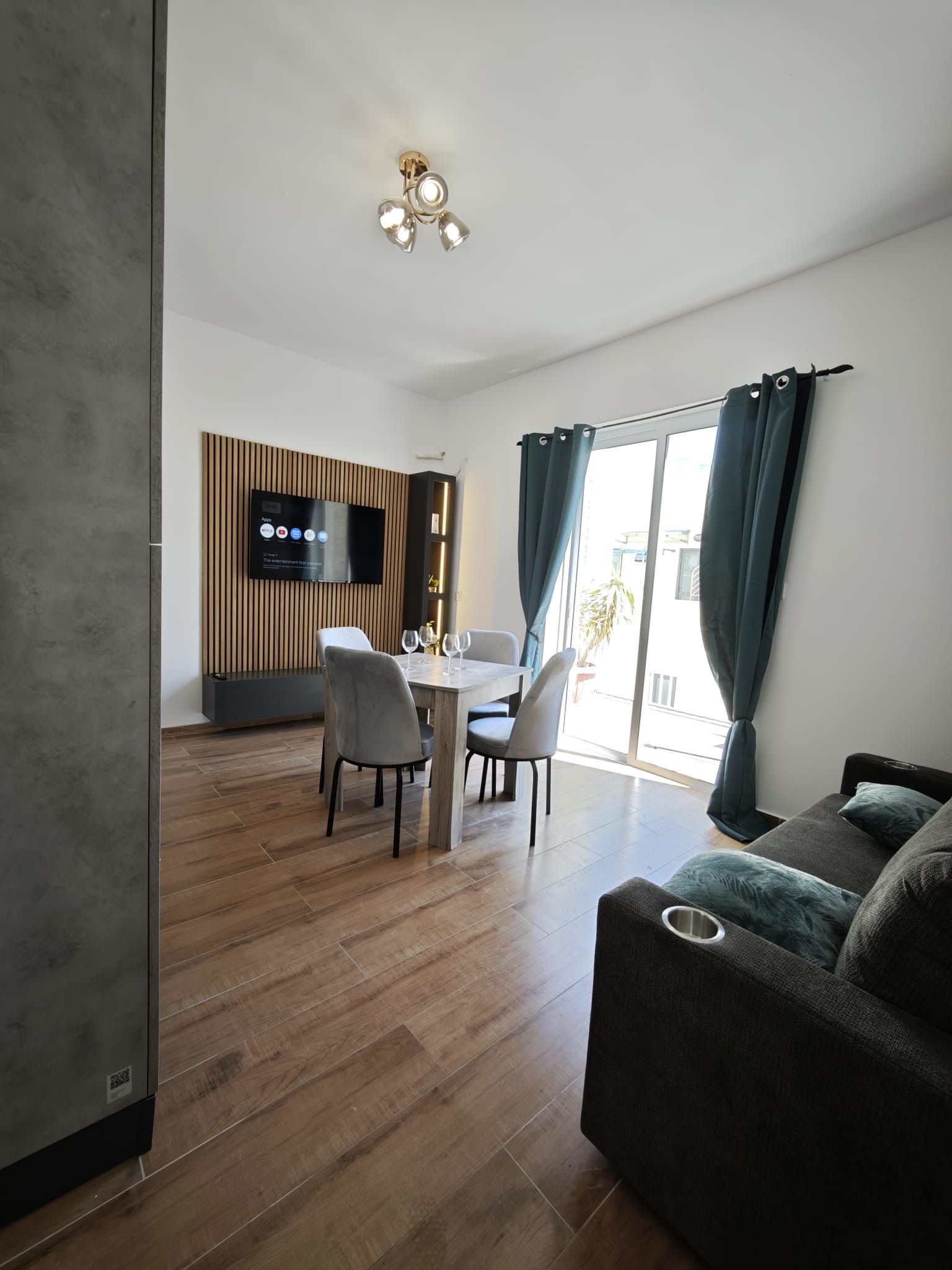 A modern living room with a dining area, a flat-screen TV mounted on a wooden panel, and a large window with curtains.
