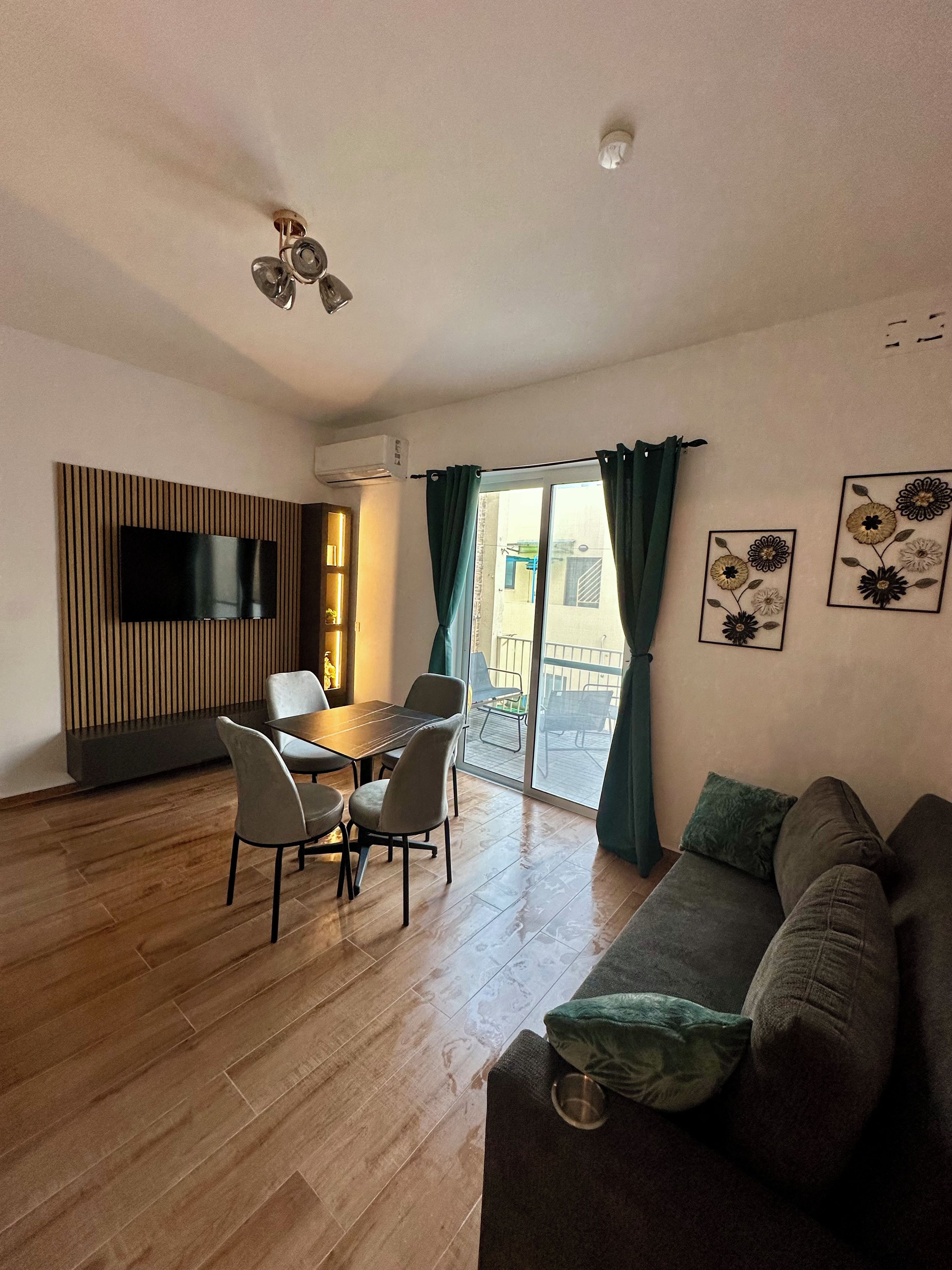 A modern living room with a flat-screen TV, a dining area with a table and chairs, a sofa, and a balcony with a view.