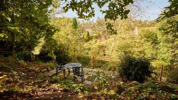 Un jardín con una mesa y sillas, rodeado de follaje denso y un muro de piedra.