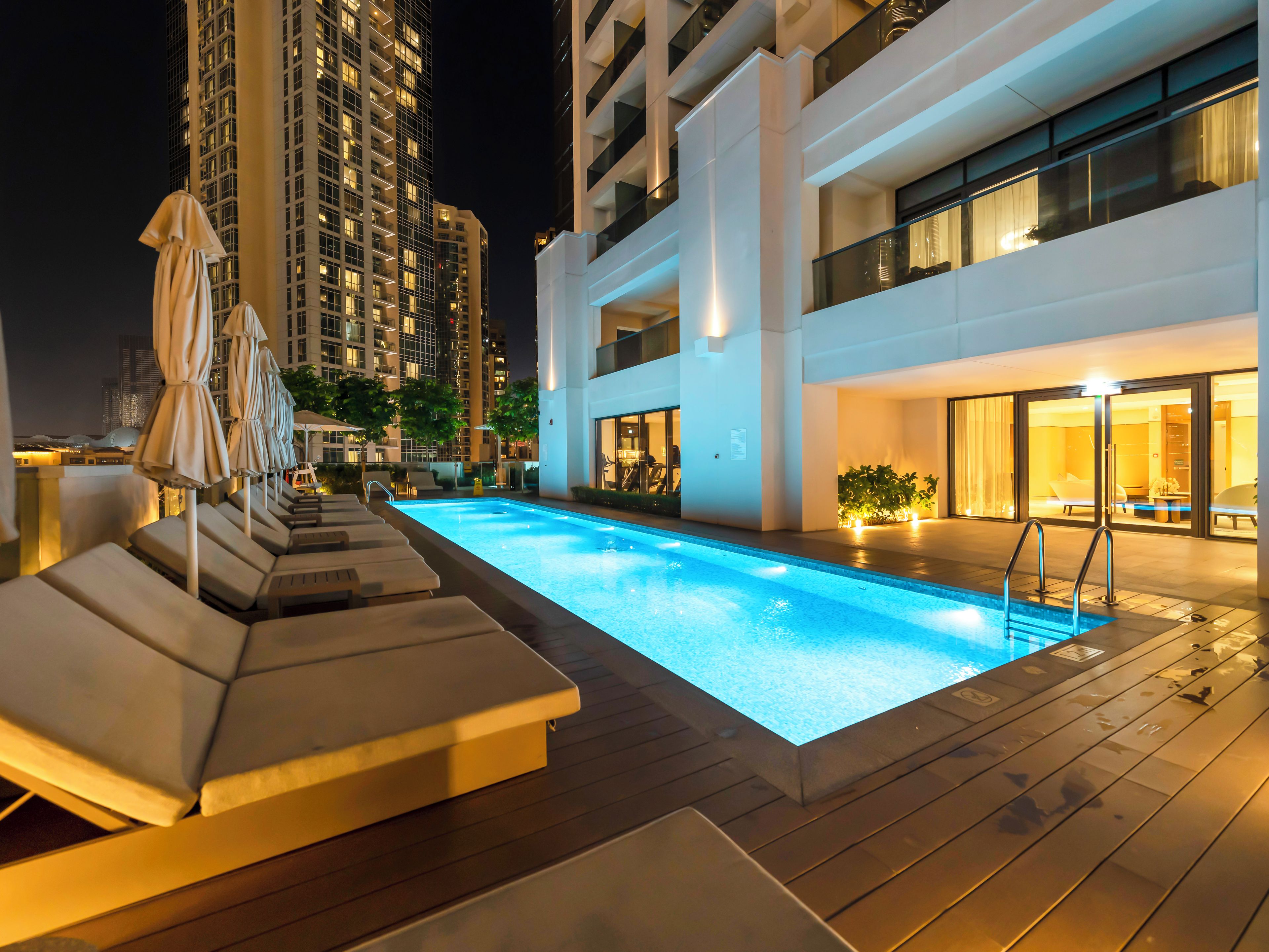 Ein Pool mit Liegestühlen und Sonnenschirmen, umgeben von modernen Gebäuden bei Nacht.