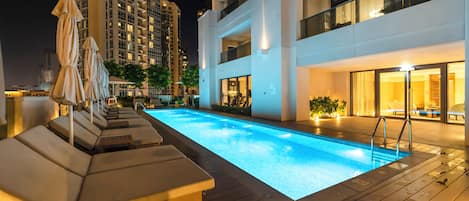 A swimming pool with lounge chairs and umbrellas, surrounded by modern buildings at night.