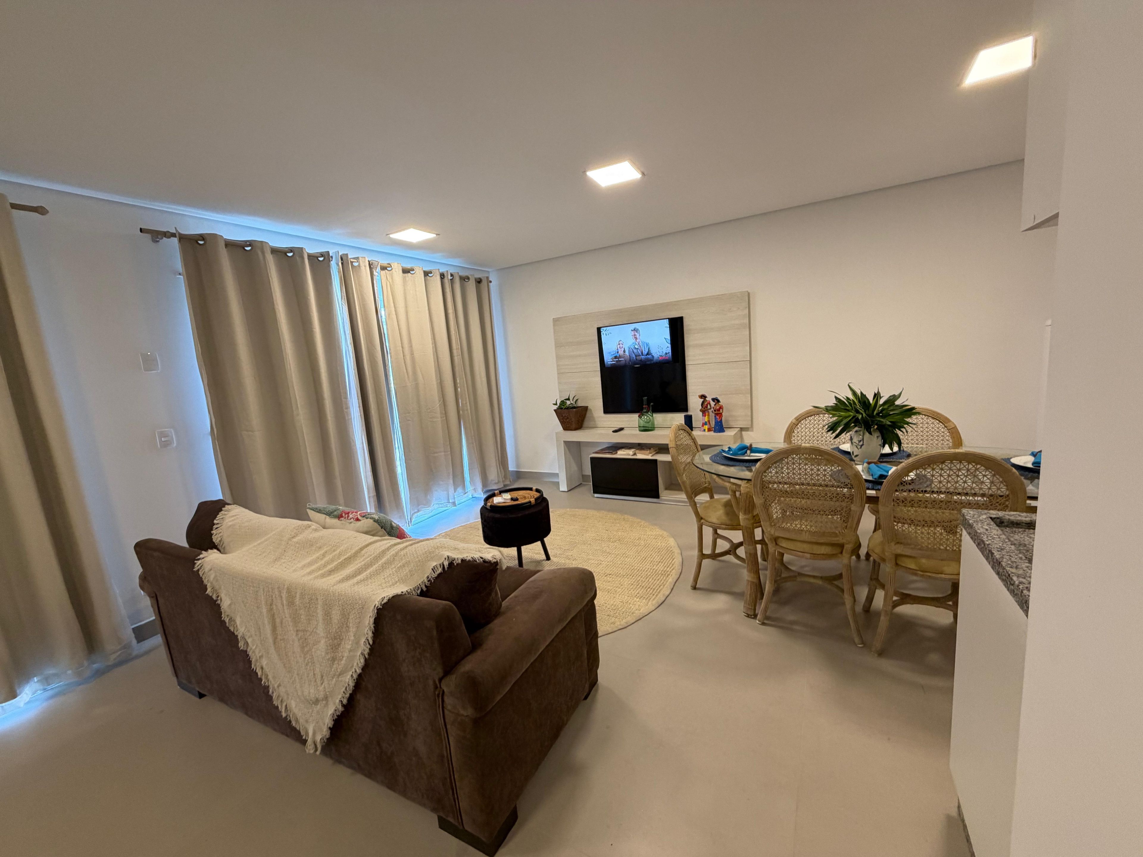 A living room with a sofa, a dining table with chairs, a TV mounted on the wall, and a small side table.