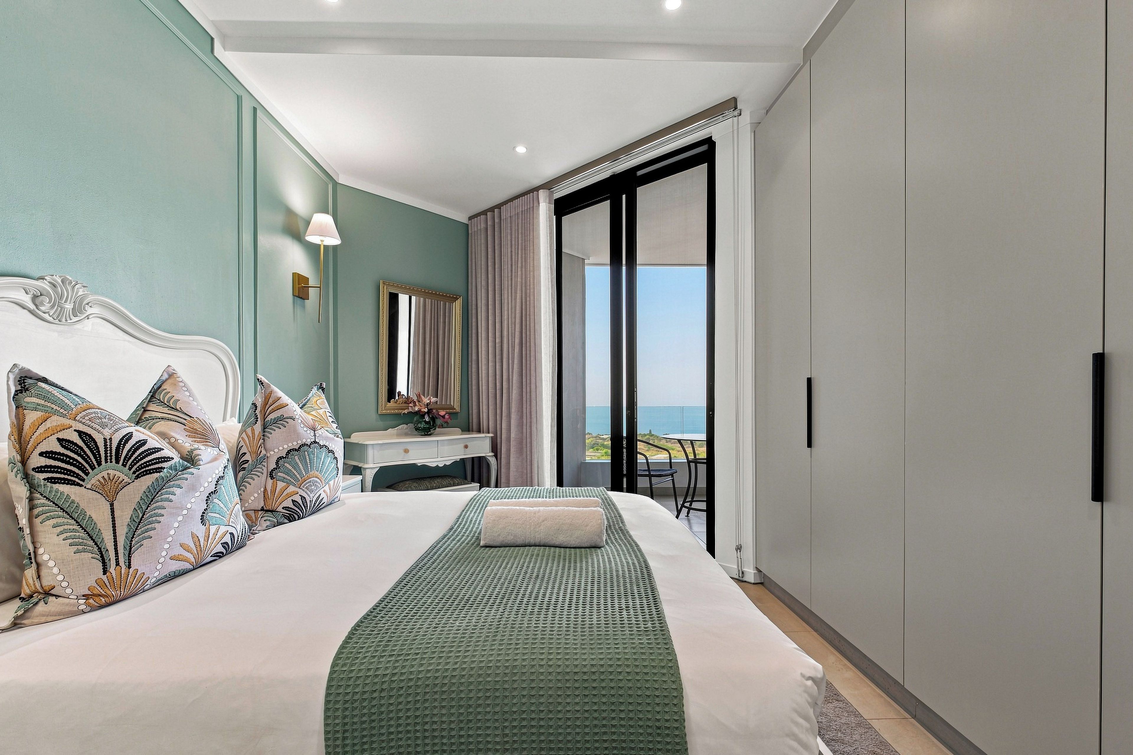 A hotel room with a large bed, a desk, a mirror, and a view of the sea.