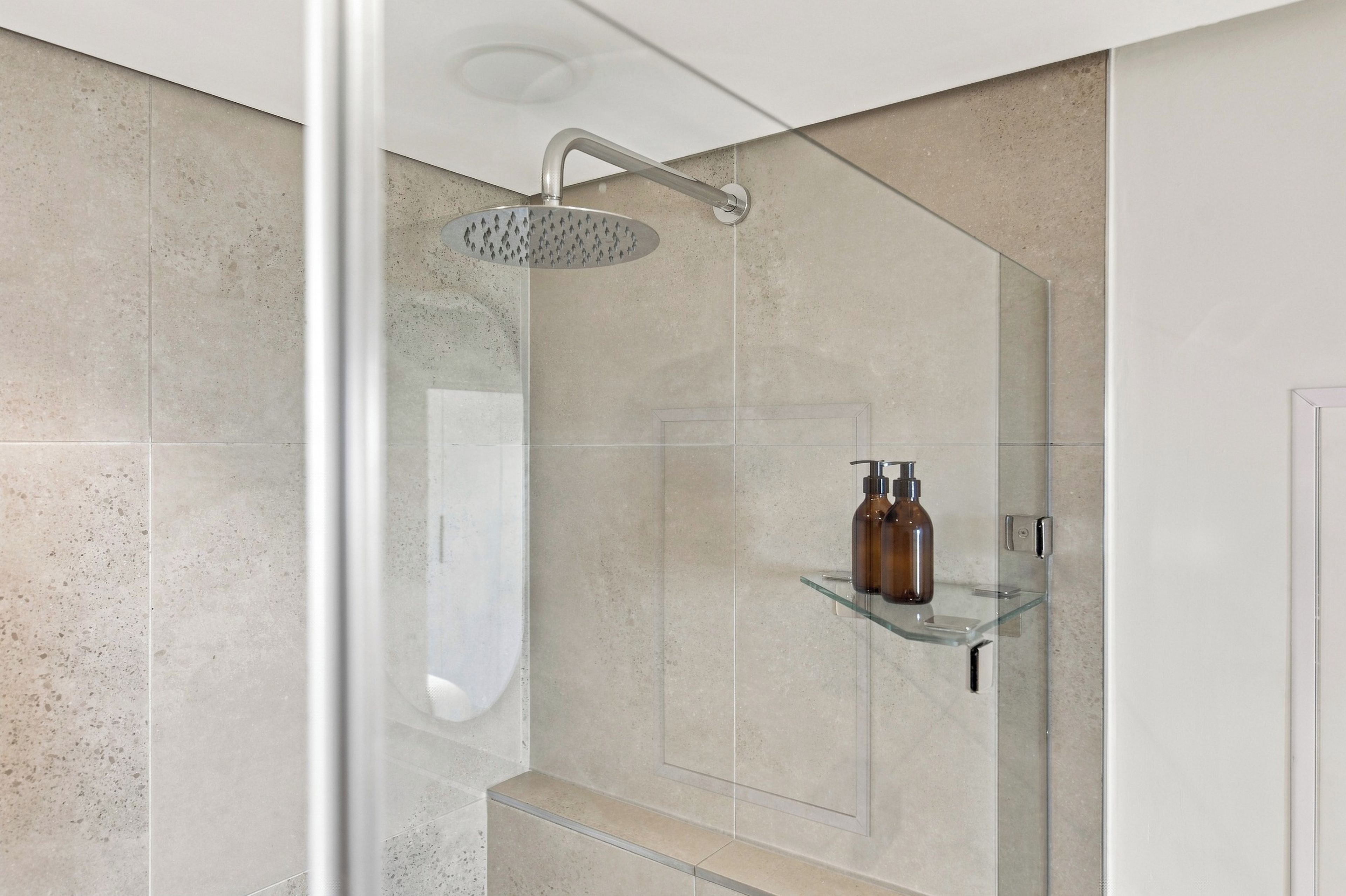Eine moderne Dusche mit Regendusche, einer Glasablage für Pflegeartikel und einem sichtbaren Toilettenpapierhalter.