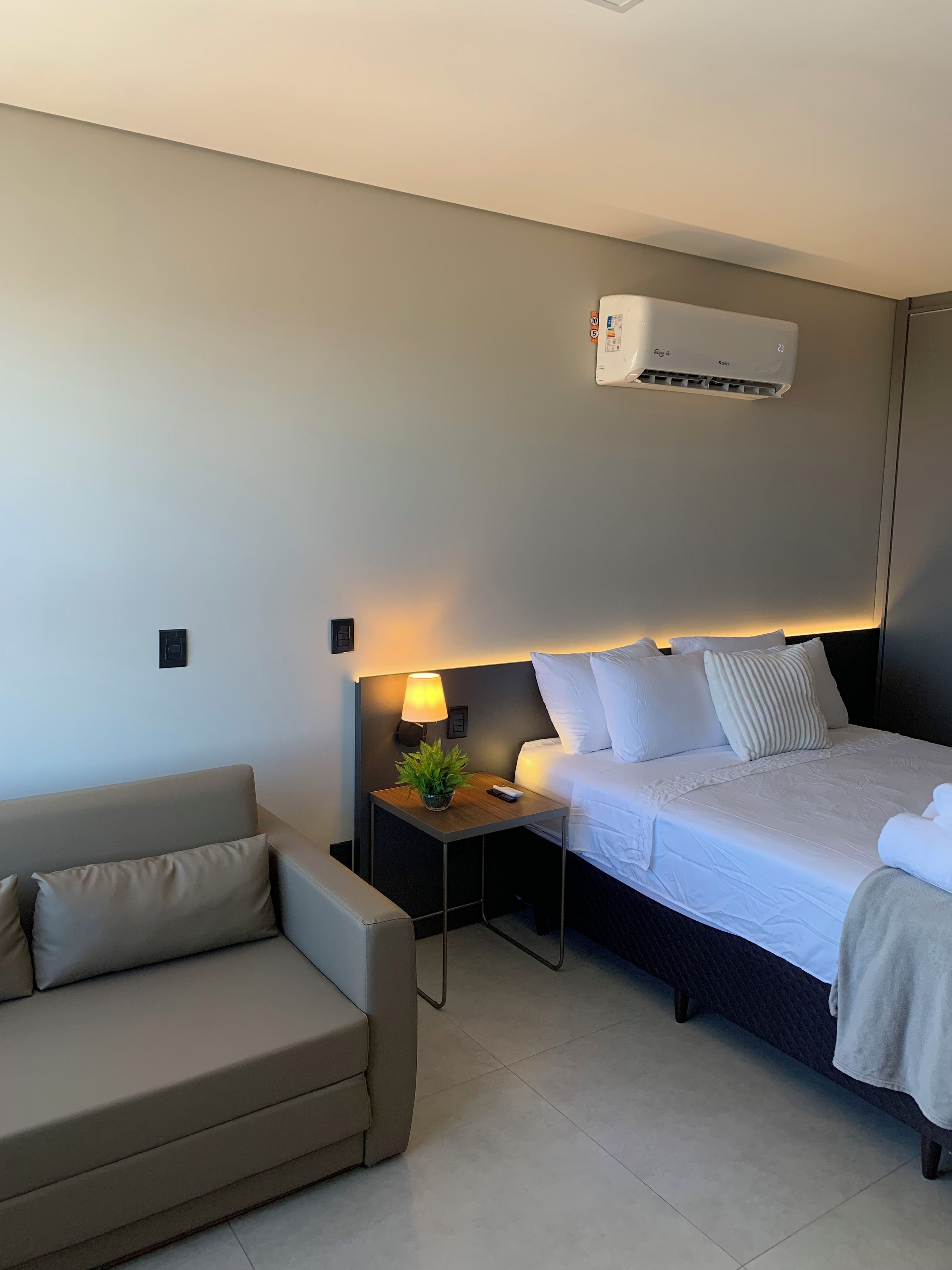 A hotel room with a bed, a bedside table with a lamp, a small potted plant, and a wall-mounted air conditioner.