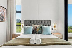 En pænt redt seng med gråt sengegavl, to sengeborde med lamper og et vindue med udsigt til grøn bevoksning.