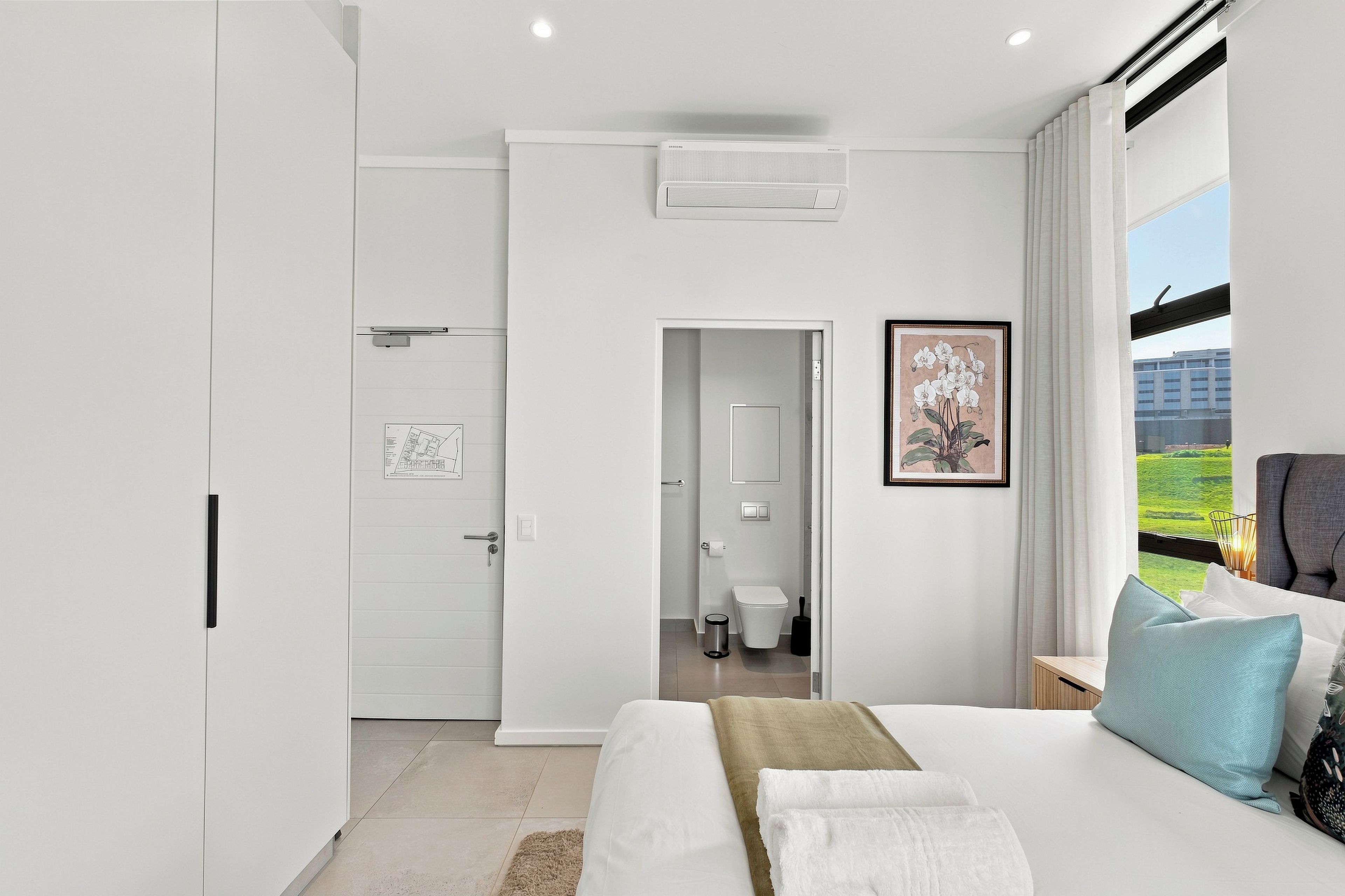 A modern bedroom with a bed, a nightstand, a window with a view, and a door leading to a bathroom.