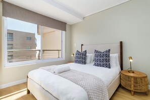 Quarto com cama grande, janela com vista para prédios e luminária de cabeceira.