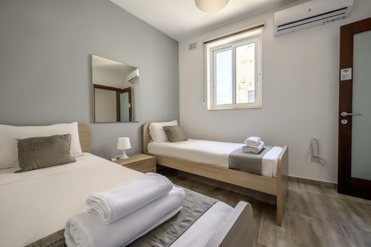 Ein Hotelzimmer mit zwei Betten, einem Fenster, einer Klimaanlage und einer Tür.
