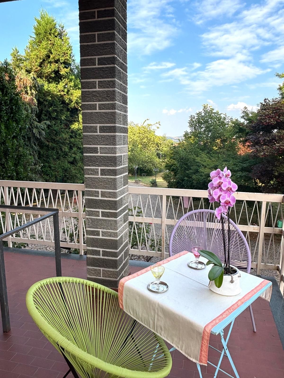 En udendørs terrasse med et bord, stole, en potte med orkidé og udsigt til træer og himlen.