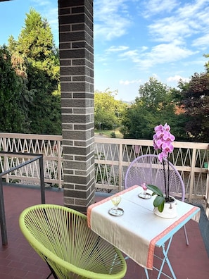 Um alpendre exterior com uma mesa e cadeiras, uma orquídea em vaso e vista para árvores e céu.