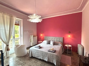 Quarto com cama, mesas de cabeceira, luminária suspensa, radiador e janela com cortinas.