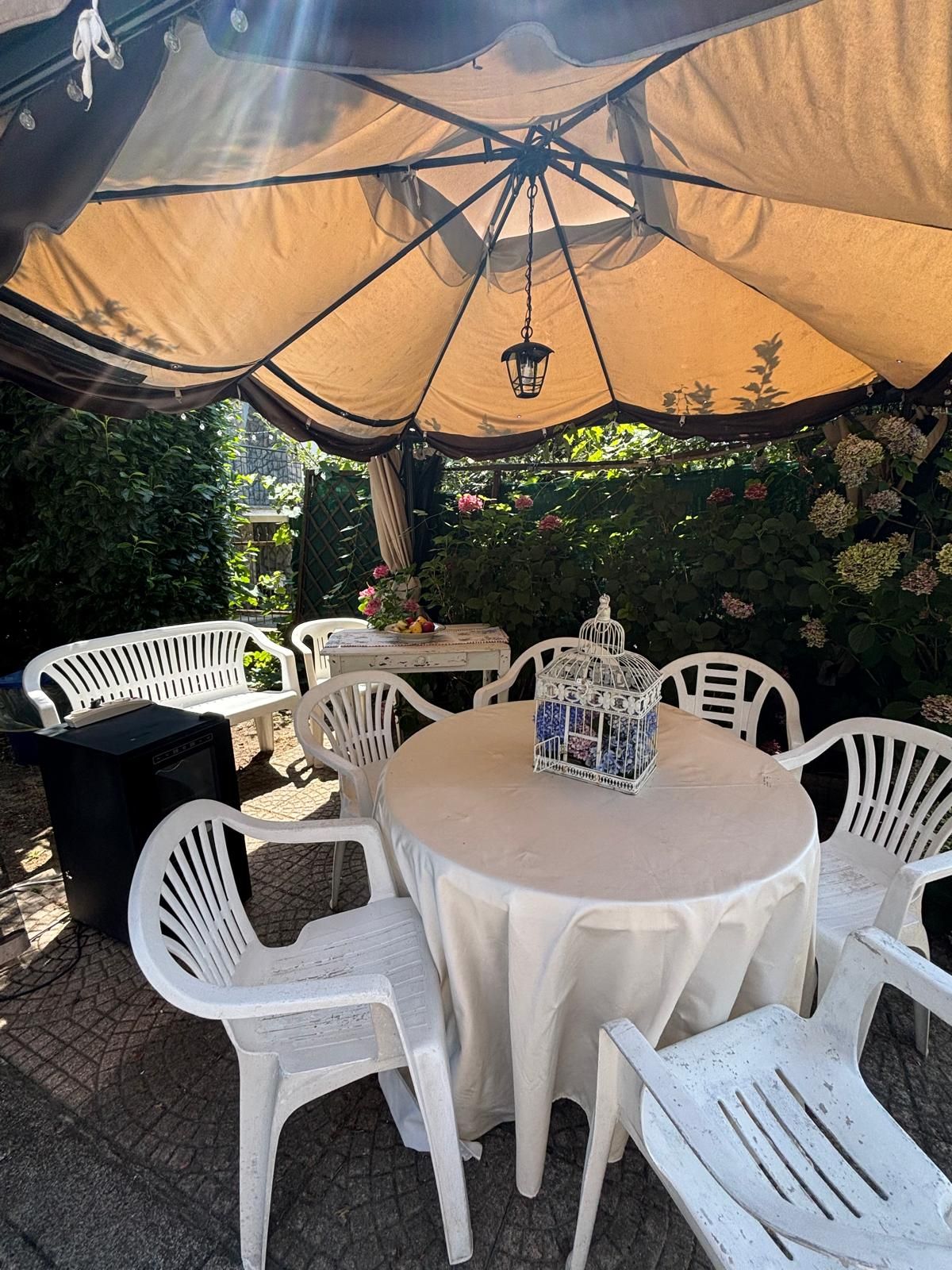 Ein Außenbereich mit einer Terrasse, einem runden Tisch und weißen Plastikstühlen, einem hängenden Laternen und einem Vogelkäfig auf dem Tisch.
