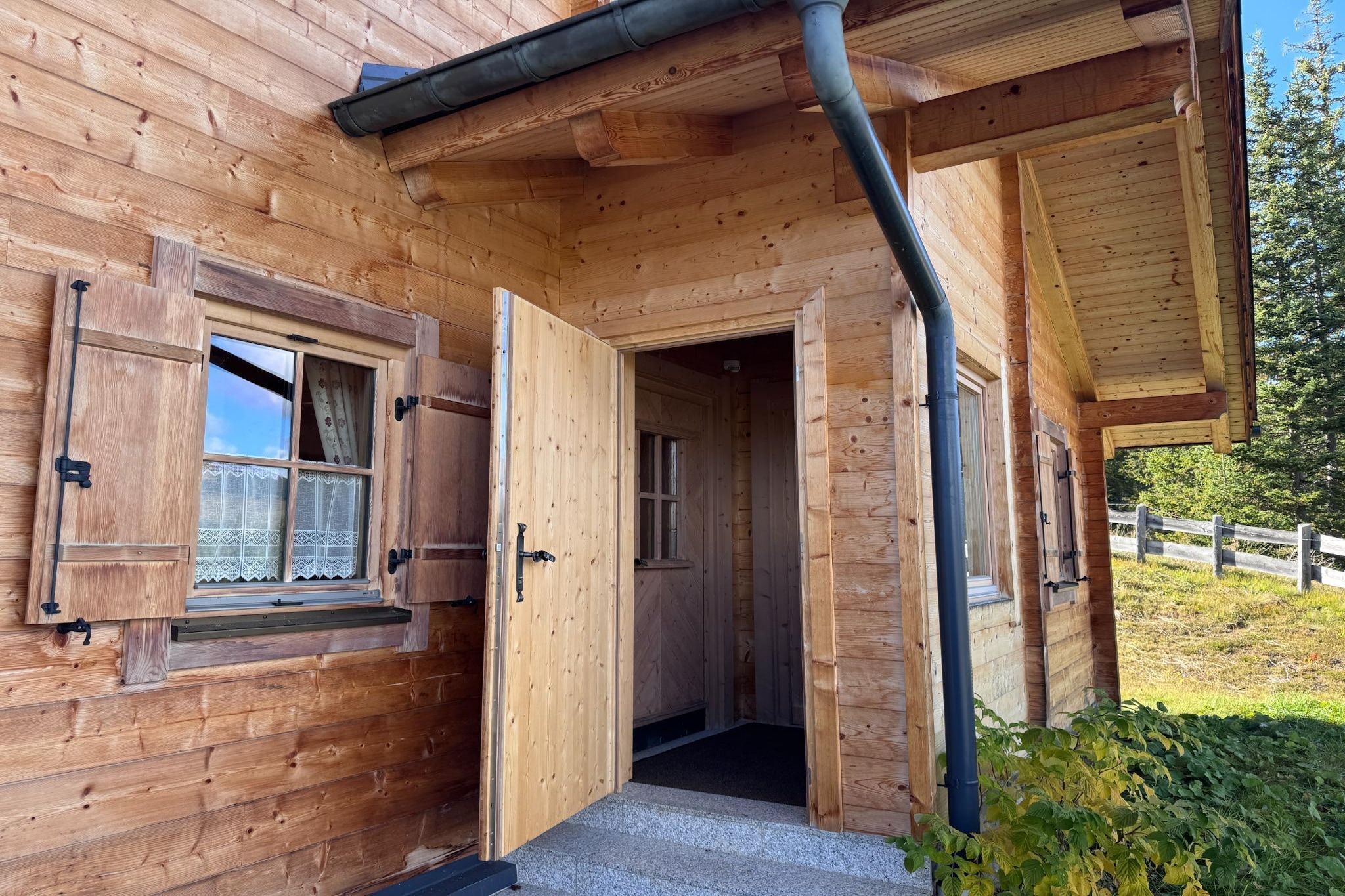 Una cabaña de madera con una puerta que da a un espacio interior, una ventana con contraventanas y un área exterior visible con una cerca.