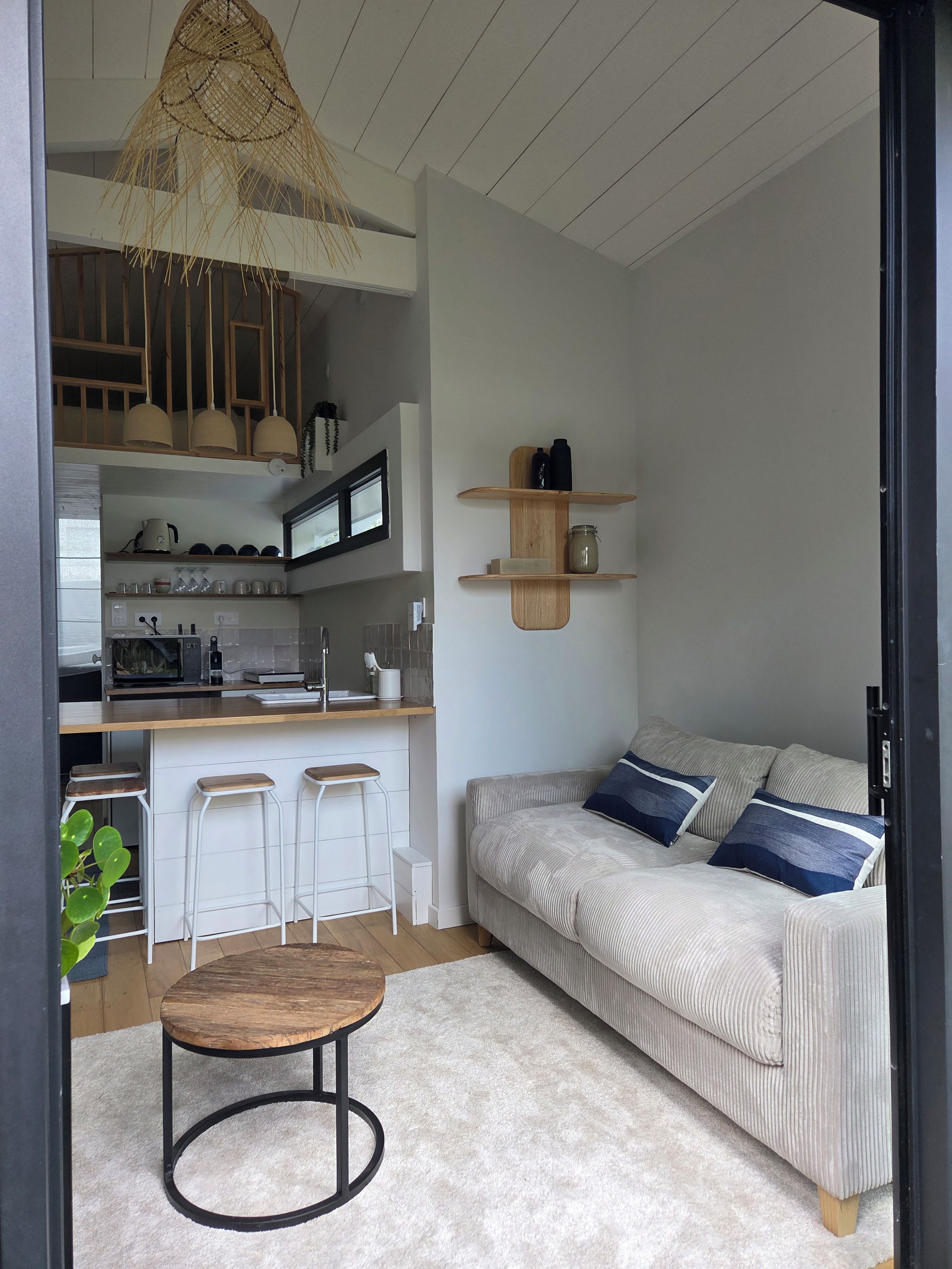 A modern living space with a kitchen area, a sofa with cushions, a round wooden table, and a hanging light fixture.