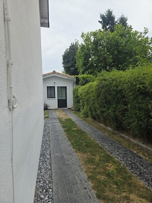 A pathway leading to a white building with a door and windows, surrounded by greenery and a gravel path.