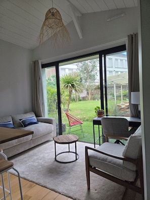 Sala de estar moderna com sofá, cadeiras, uma mesa pequena e vista para um jardim através de grandes janelas.