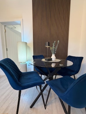 Uma sala de jantar com mesa de vidro cercada por quatro cadeiras azuis, um vaso com folhagem e uma luminária sobre a mesa.