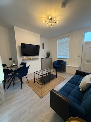 Uma sala de estar moderna com uma TV de tela plana, uma mesa de centro de vidro, um sofá azul e uma área de jantar com cadeiras.