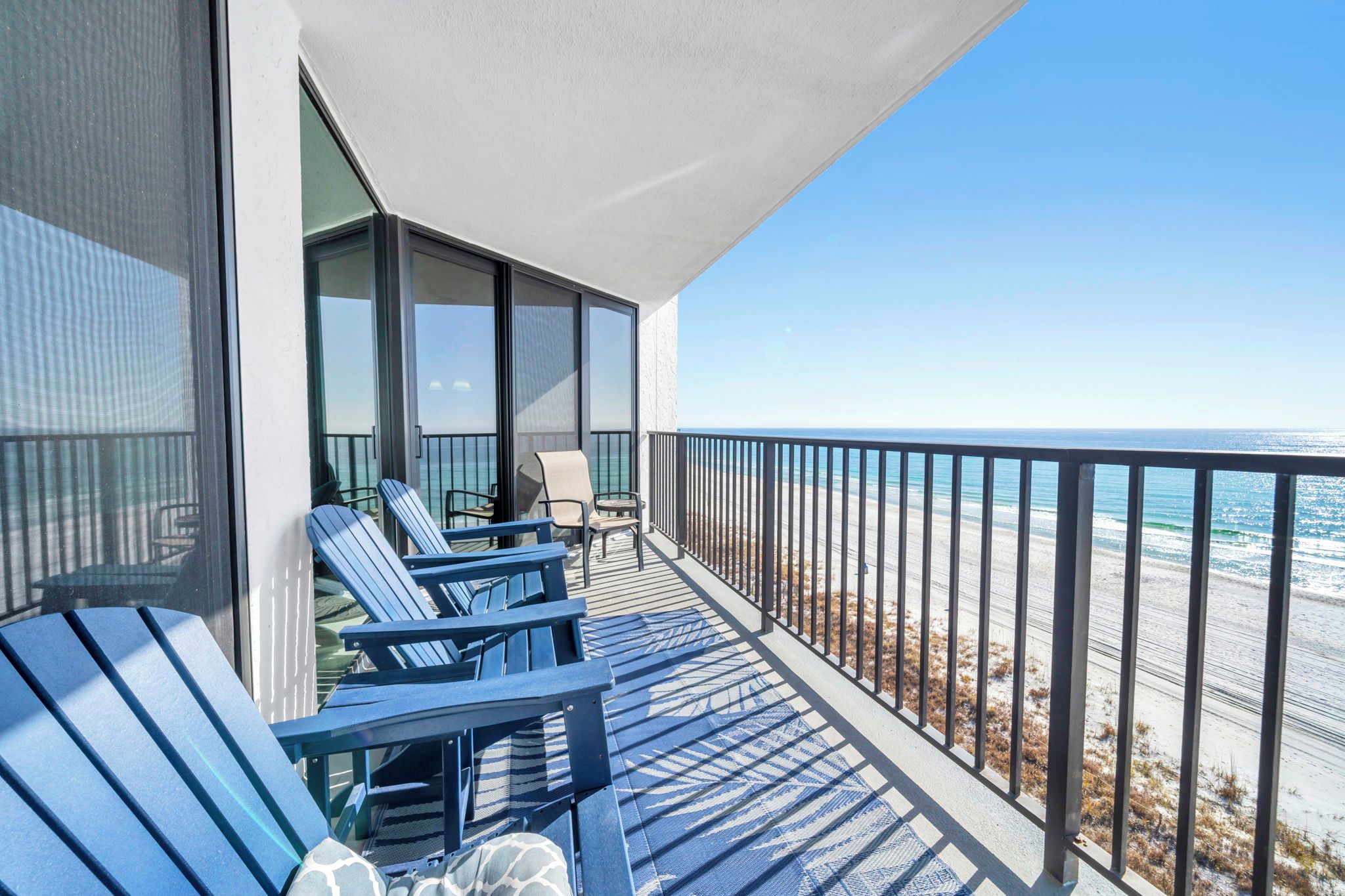 A balcony with blue Adirondack chairs, a small table, and a chair, overlooking a beach with a clear blue sky.