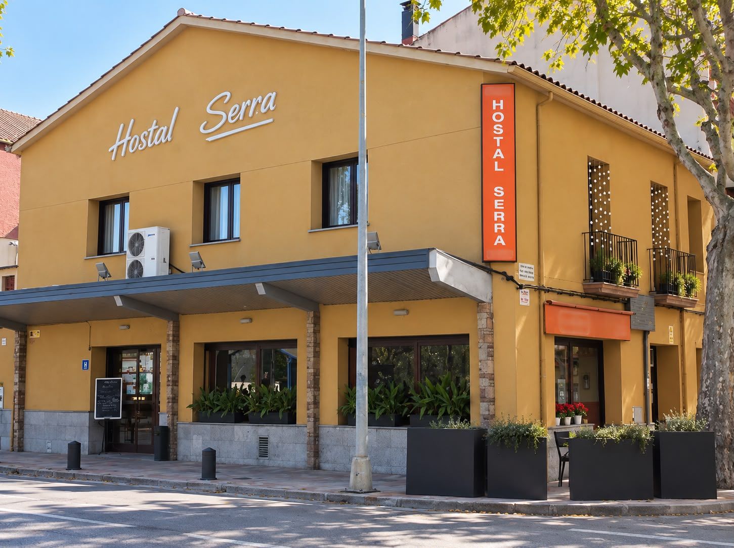 Un hostal llamado 'Hostal Serra' con fachada amarilla, unidades de aire acondicionado, entrada cubierta y zona de estar exterior con plantas.
