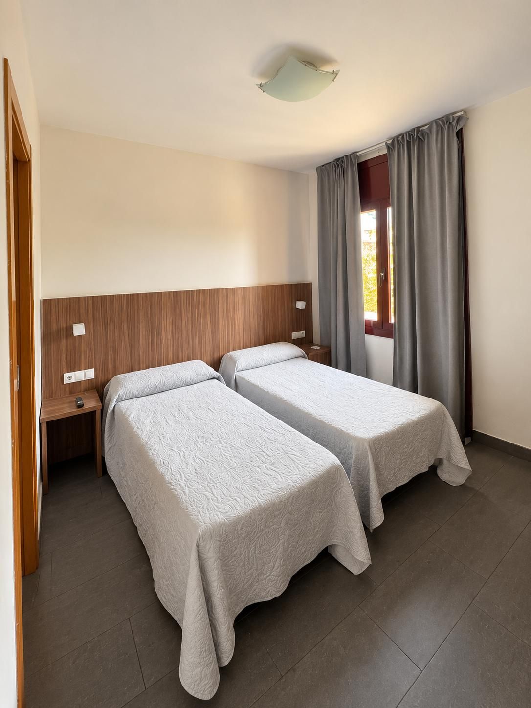 Una habitación de hotel con dos camas individuales, un cabecero de madera, una mesita de noche y un ventanal con cortinas.