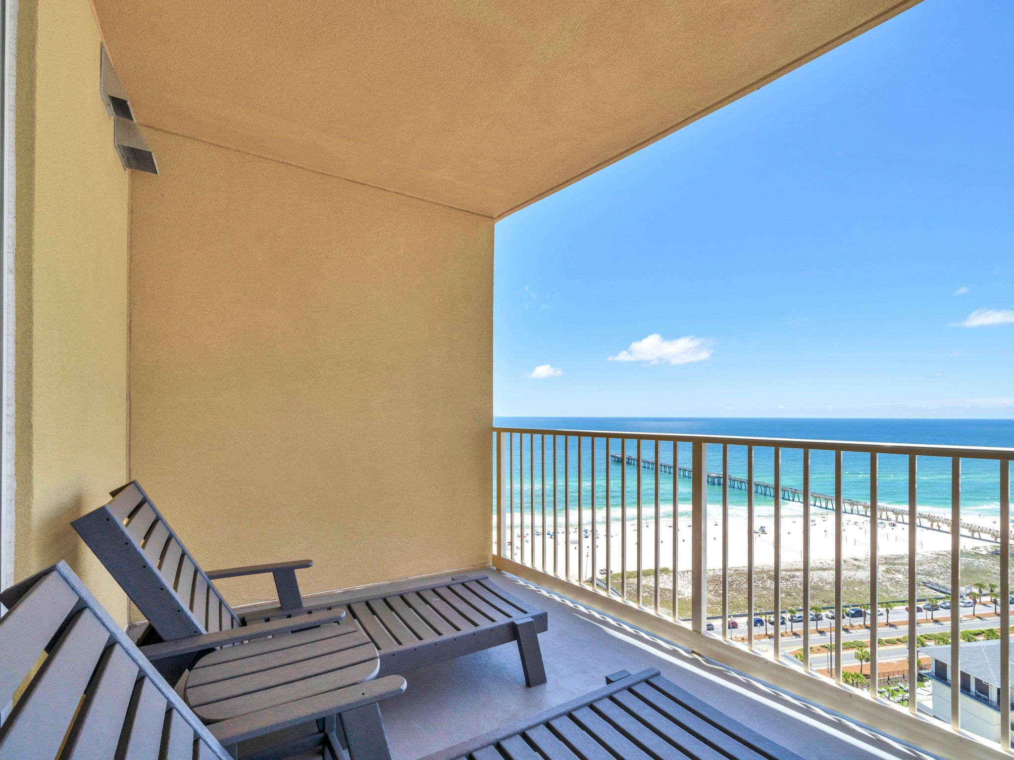 A balcony with two wooden chairs, a view of the beach, and a pier in the distance.