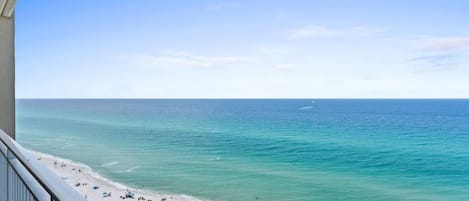 A beach view from a balcony with a clear sky, a sandy beach, and a calm sea.