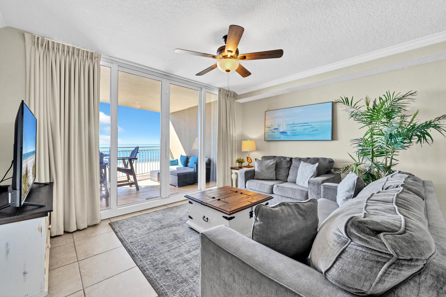 A modern living room with a grey sofa, a coffee table, a TV, and a ceiling fan. There is a sliding glass door leading to a balcony with a view of the ocean.