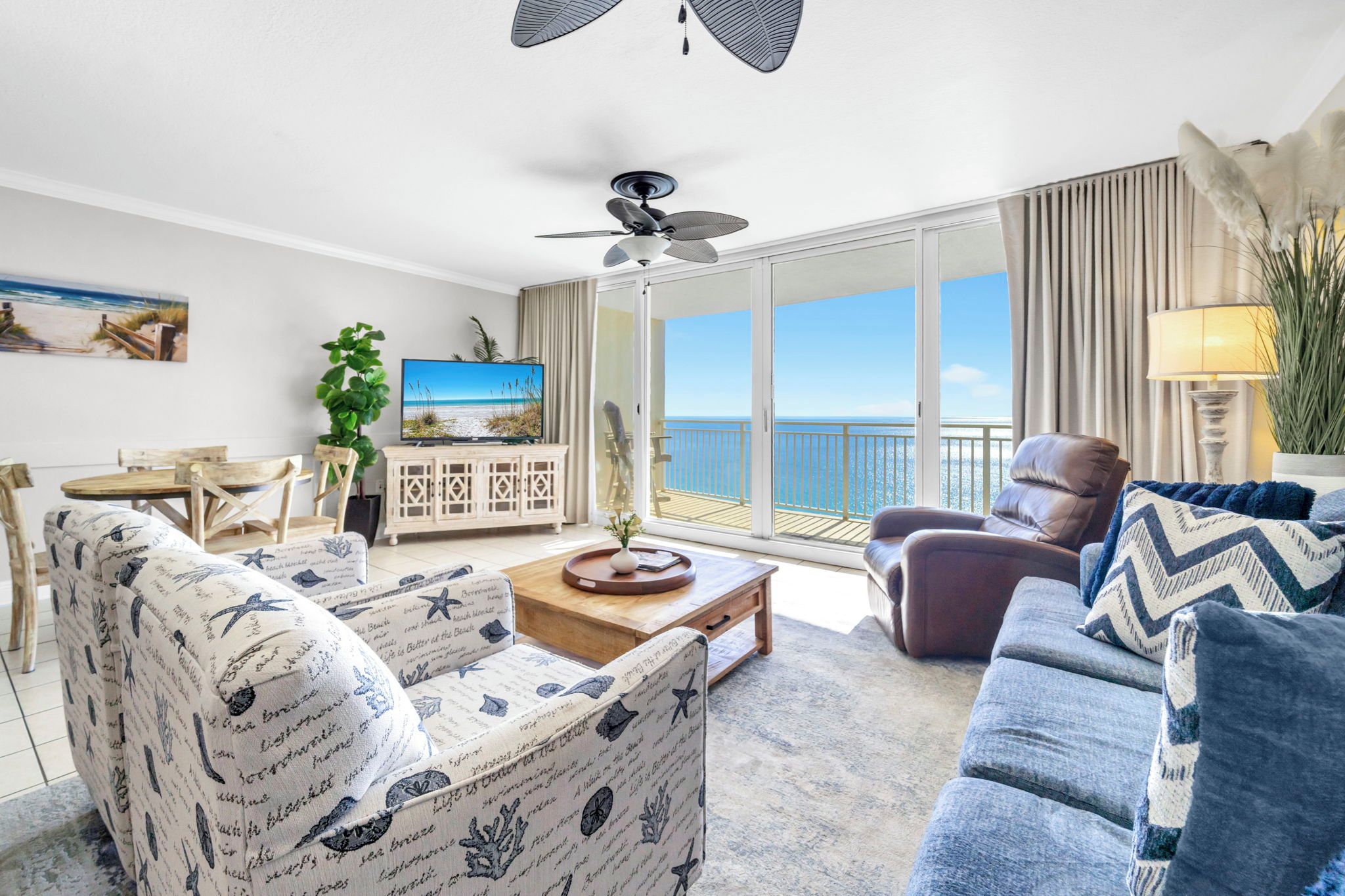 A living room with a sofa, armchair, coffee table, and TV. There is a balcony with a view of the ocean.