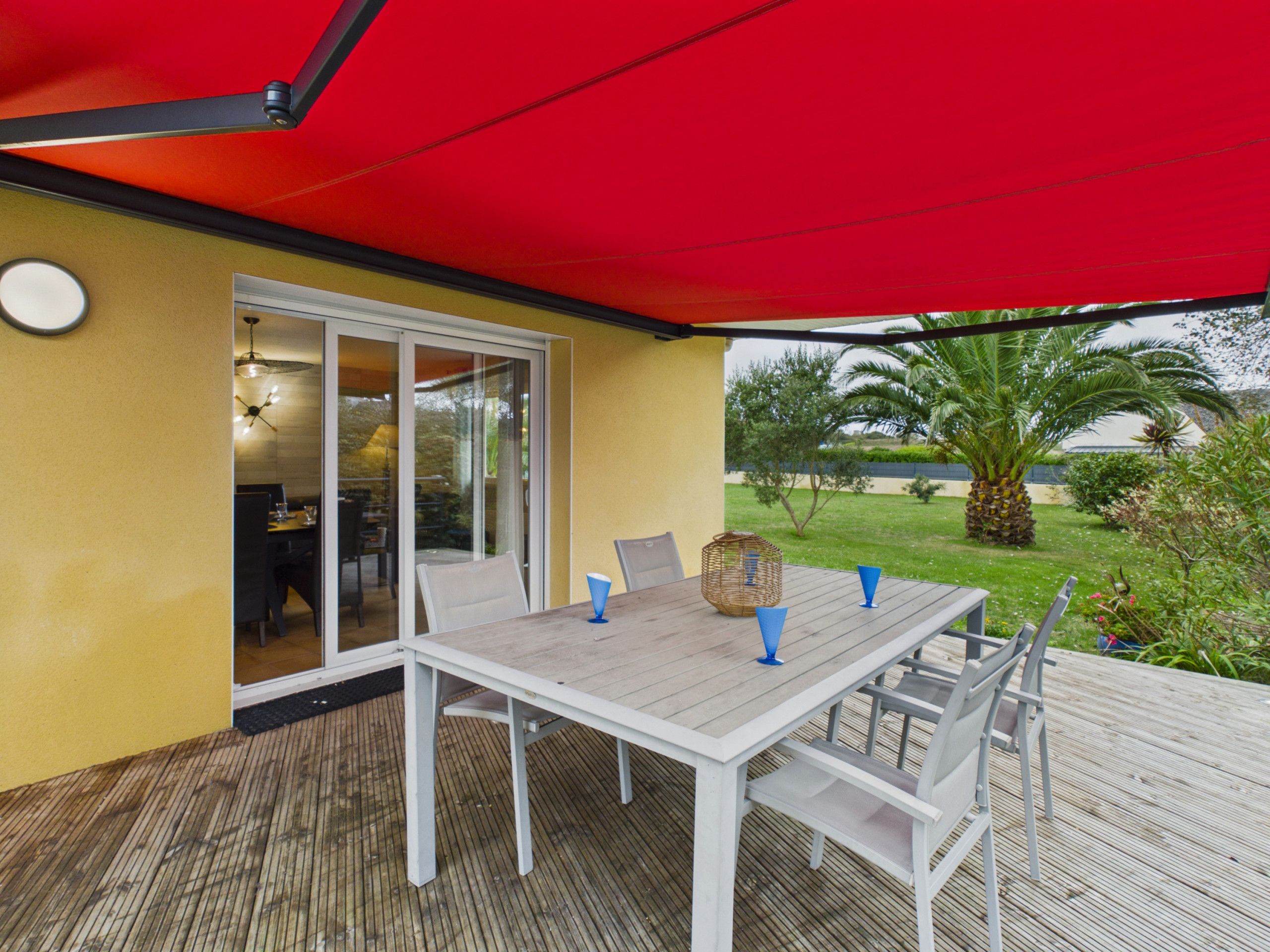 Eine Außenpatio mit einem weißen Tisch und Stühlen, einem roten Sonnensegel und Blick auf einen Garten mit Palmen.
