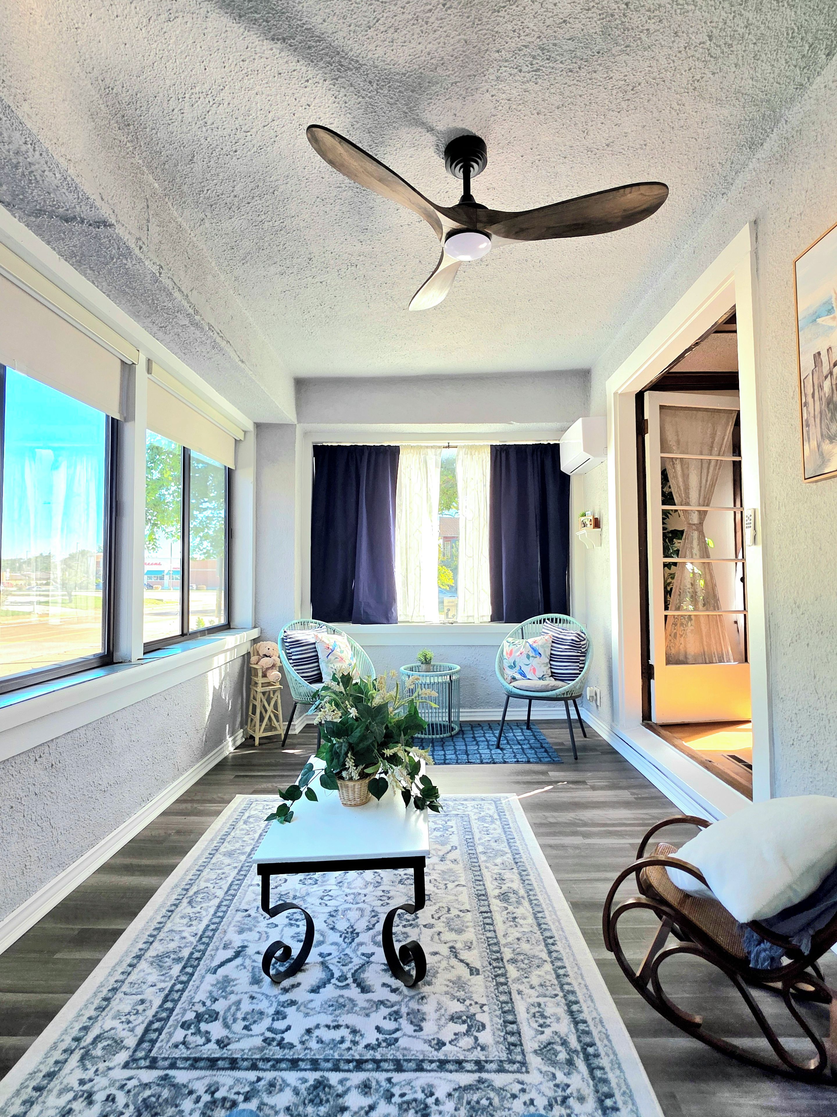 A room with a ceiling fan, large windows, a table with a plant, and two rocking chairs.