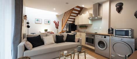 A compact living space with a sofa, a dining table, a kitchen area with appliances, and a staircase.