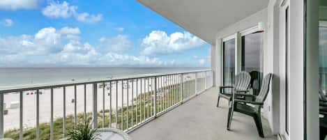 A balcony with a view of the beach and ocean, a round table with a plant, and two striped chairs.