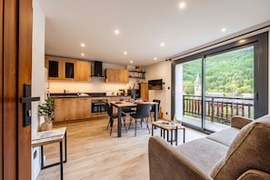 A modern kitchen with wooden cabinets, a dining area with a table and chairs, and a large sliding glass door leading to a balcony with a view of a forested area. - Perce Neige - Proche piste olympique Luc (Saint-Chaffrey)