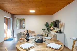 Un comedor con una mesa redonda de madera dispuesta para cuatro personas, un sofá, un televisor y una planta en maceta.