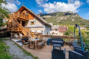 Una terraza de madera con mobiliario exterior, una parrilla y vistas a la montaña.