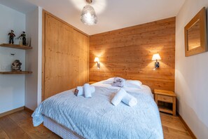 Un dormitorio con cabecera de madera, dos lámparas de noche, una cama con ropa de cama blanca y un cojín con forma de oso polar, una mesita de noche de madera y un estante con figuritas.