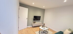 A modern living room with a flat-screen TV mounted on the wall, a white console table with decorative items, a round coffee table with a vase and remote, and a green sofa. - NEW I 3 BEDROOM HOUSE I DUDLEY I WEST MIDLANDS I POOL TABLE I FAMILY (Dudley)